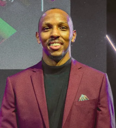 Richard Maina smiling broadly, in a burgundy blazer over a dark roll-neck, photographed from the chest up